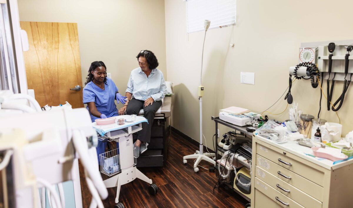 A multiracial senior woman sitting on an exam table in a doctor's office. A female doctor is sitting next to her, tending to her injured arm. The doctor is a mid adult African-American woman wearing scrubs. The patient has a wound on her arm, covered in a bandage.