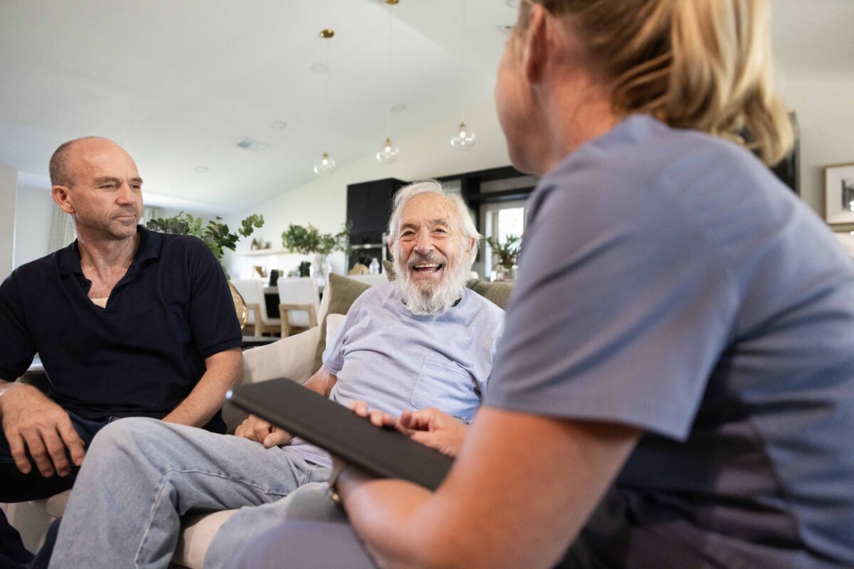 A senior gentlemen receiving health care from a nurse at home with his son present.