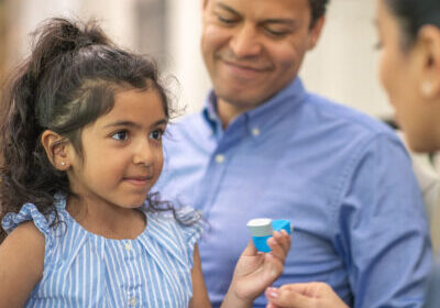 A man of Latin descent takes his daughter to see the doctor. The girl has asthma and is holding a puffer. The doctor is teaching the child how to use the medical device.