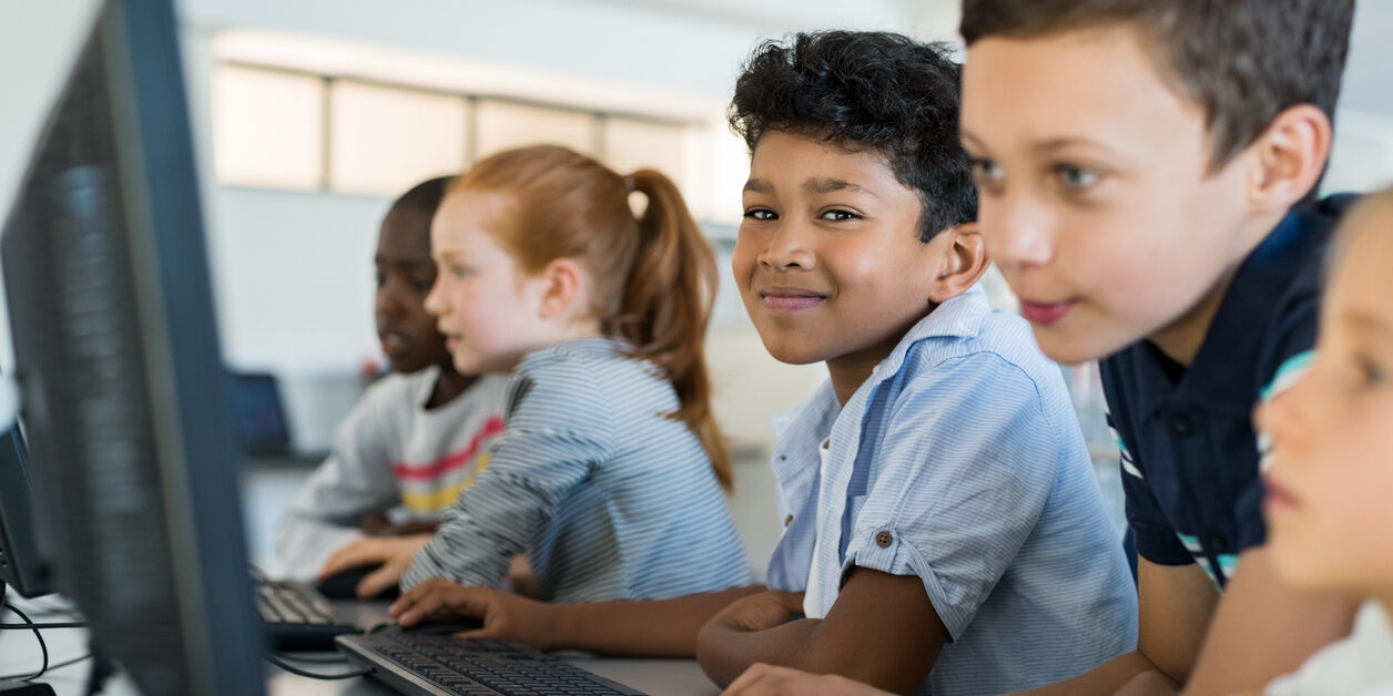 Multiethnic school kids using computer in classroom at elementary school. Portrait of arab boy looking at camera in a computer room. Smiling primary student in a row using desktop pc in class room.